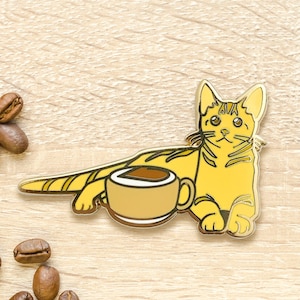 May include: Gold enamel pin of a yellow cat holding a cup of coffee. The cat is sitting on its hind legs and has a relaxed expression.