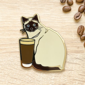 May include: A gold-tone enamel pin of a Siamese cat holding a glass of coffee. The cat has blue eyes and a white body with brown markings. The coffee is brown and has a white rim.