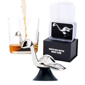 May include: A silver-colored Scotch Ness Critter whisky stone in a glass of amber liquid, with another stone in a black box. The box reads "SCOTCH NESS CRITTER WHISKY STONE". The stone is a stylized depiction of the Loch Ness Monster.