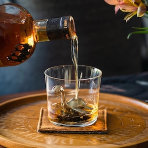 May include: A clear glass with a metal dinosaur ice mold inside is being filled with amber-colored liquid from a bottle labeled "Batch 2". The glass is sitting on a brown wooden coaster.