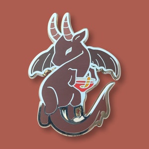 May include: A silver enamel pin of a brown, winged, cartoonish creature with horns holding a red cocktail glass. The creature has a tail that curls around its body.