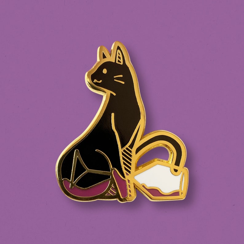 Puede incluir: Pin de esmalte dorado con un gato negro sentado en una tetera blanca y morada. El gato tiene un contorno negro simple y la tetera tiene un asa y un pico blancos.