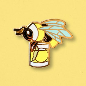 Könnte beinhalten: Goldene Emaille-Anstecknadel mit einer Biene mit gelben und schwarzen Streifen, die ihren Kopf in ein Glas Limonade taucht. Die Biene hat durchsichtige blaue Flügel.