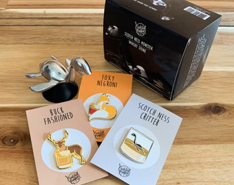 May include: Four enamel pins with different designs on white cards. The pins are themed around cocktails and feature a fox, a deer, a monster, and a glass of whiskey. The cards have the text "Foxy Negroni", "Buck Fashioned", "Scotch Ness Critter", and "Scotch Ness Monster Whisky Stone".