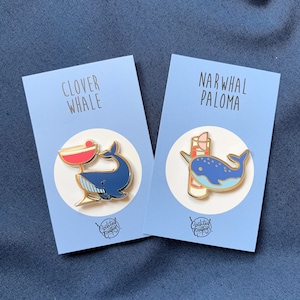 Może przedstawiać: Dwie emaliowane przypinki przedstawiające wieloryba i narwala z koktajlami. Przypinka z wielorybem ma napis "Clover Whale", a przypinka z narwalem "Narwhal Paloma". Obie przypinki mają niebiesko-białą kolorystykę i pochodzą od "Cocktail Crafts".