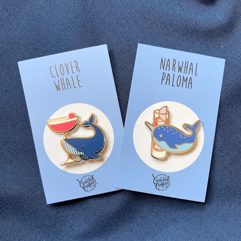 Może przedstawiać: Dwie emaliowane przypinki przedstawiające wieloryba i narwala z koktajlami. Przypinka z wielorybem ma napis "Clover Whale", a przypinka z narwalem "Narwhal Paloma". Obie przypinki mają niebiesko-białą kolorystykę i pochodzą od "Cocktail Crafts".