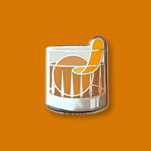 May include: Silver enamel pin of an old fashioned cocktail with an orange twist. The glass is filled with a brown liquid and the orange twist is sticking out of the top.
