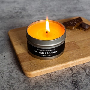 Puede incluir: Una lata de vela plateada redonda y encendida con una etiqueta negra que dice "Salted Caramel" y "NATESNECTARANDMORE.COM". La vela está sobre una tabla de cortar de madera. La vela es de 113,40 g.