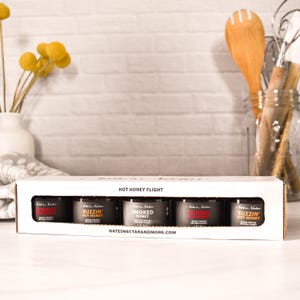 May include: A white box labeled "HOT HONEY FLIGHT" contains five small jars of flavored honey. Each jar has a black label with white text. The website address, "NATESNECTARANDMORE.COM," is printed on the box. A wooden spoon and other kitchen utensils are in the background.