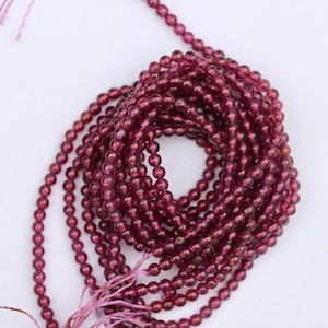 May include: A strand of deep red gemstone beads. The beads are round and have a slight shimmer. The strand is coiled and ready to be used for jewelry making.