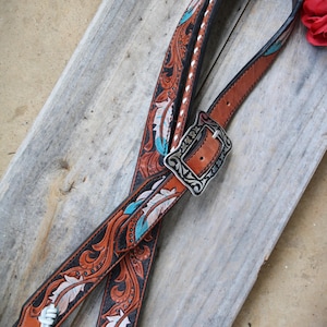 Puede incluir: Dos correas de cuero marrón con una hebilla plateada. Las correas están decoradas con un patrón floral blanco y negro y detalles turquesa. Las correas tienen borlas blancas.
