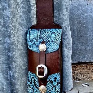 May include: A brown leather phone holster with a blue snakeskin pattern and silver buckles. The holster has a loop for a belt.