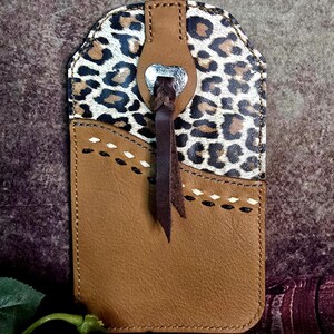 May include: A brown leather phone case with a leopard print top section. The case features a decorative heart-shaped silver metal accent and a stitched design. A dark brown leather strap hangs from the top.