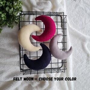 May include: Four felt crescent moon shapes in a wire basket. The moons are in cream, pink, gray, and navy blue. The text "FELT MOON - CHOOSE YOUR COLOR" is below the basket.