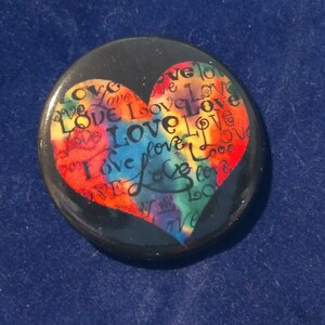 May include: A black button with a colorful heart design. The heart is filled with the word "love" written in various colors and styles.