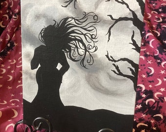 May include: A black and white painting on canvas depicting a silhouette of a woman with long flowing hair standing in front of a bare tree against a cloudy sky.