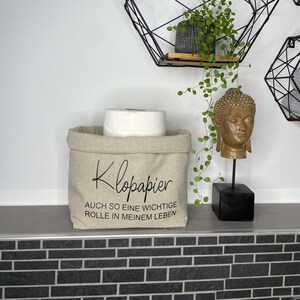Könnte beinhalten: Ein beigefarbener Stoffkorb mit dem Text "Klopapier auch so eine wichtige Rolle in meinem Leben" in schwarzer Schrift. Der Korb ist mit einer Rolle Toilettenpapier gefüllt und steht auf einem Regal mit einem schwarzen geometrischen Regal dahinter.