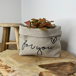 Könnte beinhalten: Ein beiger Canvas-Pflanzer mit dem Text "for you" und einem Herzsymbol. Der Pflanzer ist mit einer grün-roten Pflanze gefüllt.