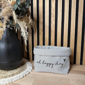 Könnte beinhalten: Ein grauer und weiß gestreifter Stoffaufbewahrungskorb mit dem Text "oh happy day" und einem kleinen gestickten Herz auf der Vorderseite.