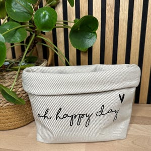 Könnte beinhalten: Ein weißer Canvas-Aufbewahrungskorb mit schwarzem Text, der "oh happy day" lautet und ein kleines schwarzes Herz. Der Korb steht auf einer Holzoberfläche.