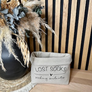 Op de afbeelding: Een beige stoffen opbergmand met de tekst "LOST SOCKS seeking soulmates" en een klein hartje.