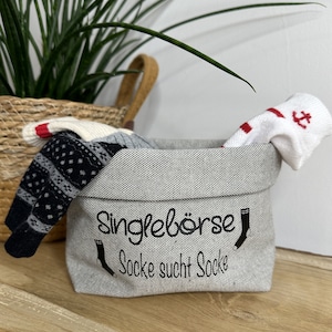 Könnte beinhalten: Ein grauer Stoffaufbewahrungskorb mit dem Text "Singlebörse Socke sucht Socke" und einer schwarzen Sockengrafik. Der Korb ist mit Socken gefüllt, darunter eine weiße Socke mit einem roten Anker und eine schwarz-weiß gemusterte Socke.