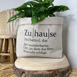 Könnte beinhalten: Ein beiger Canvas-Pflanztopf mit dem Text "Zuhause [tsu'haʊzə], das der wunderbarste Ort, an dem das Herz Wurzeln schlägt." Eine grüne Pflanze mit großen Blättern befindet sich im Topf.