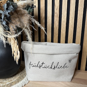 Könnte beinhalten: Eine beige Stofftasche mit weißem Streifenmuster und dem Schriftzug "frühstückliebe" in Schwarz mit einem kleinen Herzsymbol.