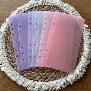 Könnte beinhalten: Fünf transparente Plastik-Seitenschützer in verschiedenen Pastellfarben, darunter Lila, Rosa und Weiß. Die Seitenschützer haben ein Lochstanzdesign für die Verwendung in einem Ordner oder Planer.