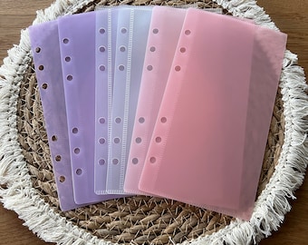 May include: Five clear plastic page protectors in different pastel colours, including purple, pink, and white. The protectors have a hole punch design for use in a binder or planner.