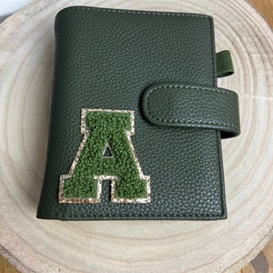 May include: A dark green leather journal with a textured finish. The journal features a green, fuzzy letter "A" patch with gold glitter trim. It has a strap closure and an elastic band. The journal is on a light wood surface.