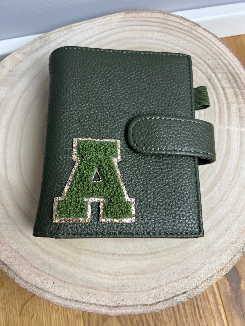 May include: A dark green leather journal with a textured finish. The journal features a green, fuzzy letter "A" patch with gold glitter trim. It has a strap closure and an elastic band. The journal is on a light wood surface.