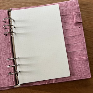 Könnte beinhalten: Ein rosafarbener Leder-Planer mit einer offenen weißen Seite mit Punkten. Der Planer hat ein Ringbindungssystem und eine Tasche auf der rechten Seite.