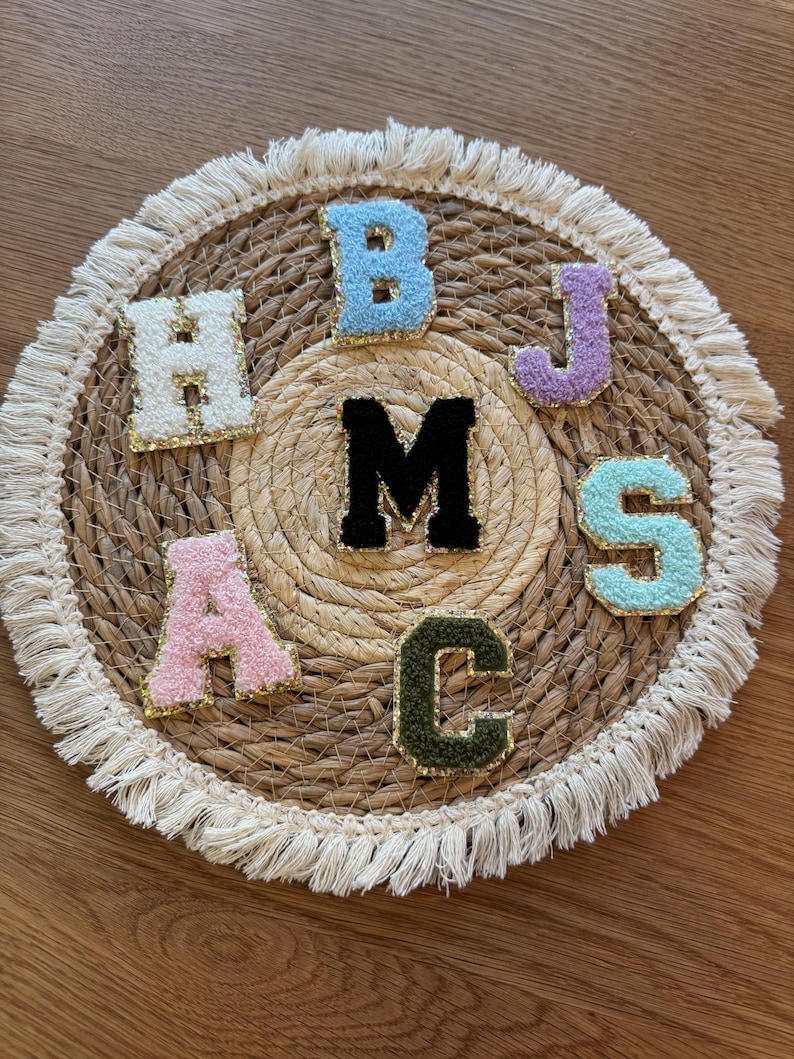 May include: A selection of embroidered fabric letters in various colours, including H, B, J, S, A, C, and M, displayed on a woven placemat. Each letter has gold trim, offering a decorative and personalised touch.