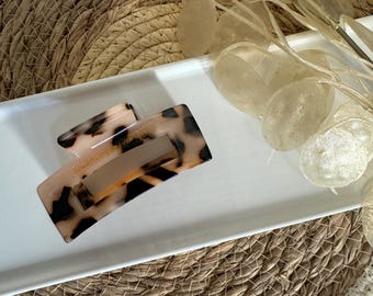 May include: A large tortoiseshell hair claw with a brown and white pattern. The claw is on a white tray with a woven straw mat underneath.