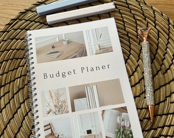 May include: A white spiral bound budget planner with the text "Budget Planner" on the cover. The cover features photos of a desk, a vase with dried flowers, a book, and a chair.