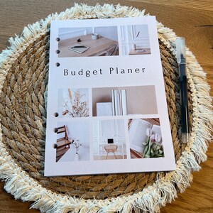 Könnte beinhalten: Ein weißer Budgetplaner mit dem Text "Budget Planner" in Schwarz. Der Planer hat auf der Vorderseite eine Collage aus Bildern, darunter ein Schreibtisch mit Laptop, ein Buch und eine Vase mit Blumen.