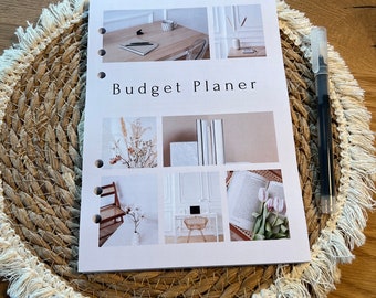 Op de afbeelding: Een witte budgetplanner met de tekst "Budget Planner" in zwart. De planner heeft een collage van afbeeldingen op de voorkant, waaronder een bureau met een laptop, een boek en een vaas met bloemen.