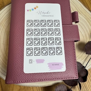 Könnte beinhalten: Ein weißer Aufkleber mit einem Wäsche-Challenge-Tracker. Der Aufkleber hat 24 Waschmaschinen-Symbole und einen Platz, um die Gesamtzahl der Ladungen zu notieren. Der Aufkleber befindet sich auf einem rosafarbenen Leder-Planer.