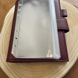 Könnte beinhalten: Ein weinroter Lederplaner mit einem transparenten Kunststoffeinsatz. Der Planer hat eine strukturierte Oberfläche und einen Schnappverschluss. Der transparente Einsatz hat mehrere Taschen zum Organisieren von Papieren und Notizen. Der Planer liegt auf einer Holzoberfläche.