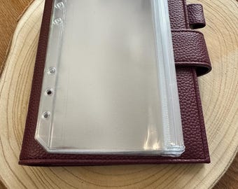 Op de afbeelding: Een bordeauxrode leren planner met een doorzichtige plastic inleg. De planner heeft een getextureerde afwerking en een druksluiting. De doorzichtige inleg heeft meerdere vakken voor het organiseren van papieren en notities. De planner ligt op een houten oppervlak.