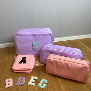 Könnte beinhalten: Vier violette und rosafarbene Reißverschlusstaschen mit verschiedenen Buchstaben auf der Vorderseite. Die Buchstaben sind A, B, D, E, F und G. Die Taschen befinden sich auf einer Holzoberfläche.