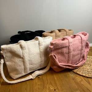 Könnte beinhalten: Vier flauschige Tragetaschen in verschiedenen Farben: Weiß, Schwarz, Beige und Rosa. Die Taschen haben lange Riemen und sind aus einem weichen, plüschigen Material gefertigt.