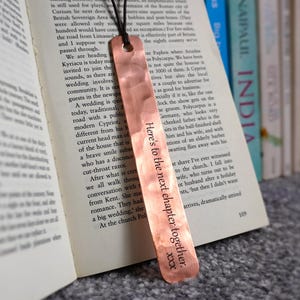 Puede incluir: Un marcapáginas de cobre con el texto "Here's to the next chapter together. xxx" cuelga de un libro. El marcapáginas es rectangular con un cordón negro. Hay libros en el fondo.