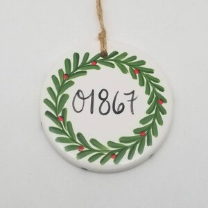 May include: White ceramic Christmas ornament with a green wreath and red berries. The number 01867 is written in black inside the wreath.