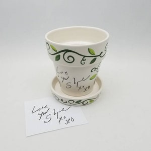May include: White ceramic flower pot with green vine design and the words "Love you S" written on it. The pot has a matching saucer. A white card with the same message is next to the pot.
