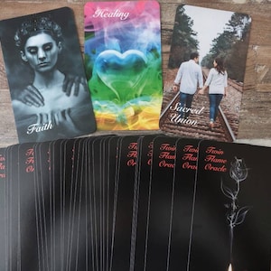May include: A deck of Twin Flame Oracle cards with three cards on top. The top cards read "Faith", "Healing", and "Sacred Union". The deck has a black background with a rose illustration and the words "Twin Flame Oracle".