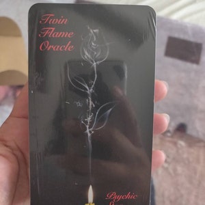 May include: A black Twin Flame Oracle card featuring a white floral design and a lit candle. The words "Twin Flame Oracle" are in red script at the top, and "Psychic Star" is in red script at the bottom.