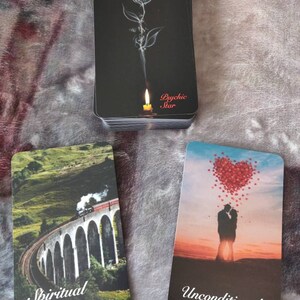 May include: A deck of Twin Flame Oracle cards with a black background and a rose design. Two other cards show a train on a bridge and a couple under a heart-shaped cloud. The cards are for spiritual guidance.