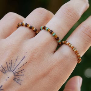 Könnte beinhalten: Drei Perlenringe in Braun- und Türkistönen werden an Fingern getragen. Die Ringe bestehen aus kleinen, farbigen Perlen. Eine Hand mit einem Sonnenaufgang-Tattoo ist im Vordergrund, mit einem verschwommenen grünen Hintergrund.